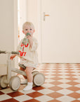 A toddler on a beige balance bike drinks from a green mushie Soft Spout Sippy Cup, wearing a light shirt with red text and white socks. The checkered red-and-white floor leads to a white door and wall in the background.
