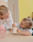 A young toddler with light hair in pigtails smiles at a table while reaching into a mushie Snack Cup. An adult with blonde hair in a ponytail smiles at the child, and a pink water bottle sits on the table.