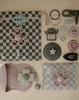 A flat lay of pastel baby accessories features two mushie Water Resistant Wet Bags for diaper bag storage, a floral bib, teething toys, pacifiers, and green, purple, and blue silicone items on a neutral background.