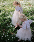 Two young children in pastel fairy costumes wander through a field of white wildflowers. The child in front carries a mushie Silicone Baby Food Container, while the other follows, both surrounded by greenery and blooms.