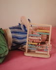 A young child in a blue striped sweater and green pants lies on a pink surface, holding up the Mushie Wooden Abacus—a Montessori counting toy with colorful pastel beads.