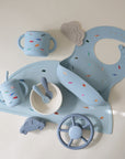 A mushie Silicone Baby Bib in light blue is shown among matching baby feeding items and toys with colorful shapes, all neatly arranged on a white background.
