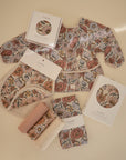 Floral-patterned baby items in soft pink, beige, and white—bib, swaddle cloths, organic cotton long-sleeve top, and two boxed sets—are displayed with packaged Mushie Changing Pad Liners on a cream surface.