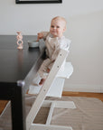 A baby in striped pajamas sits in a white high chair at a black table, holding a spoon and bowl. The floor is protected by the Mushie Splat Mat, and there's a pink candle holder on the table. The baby looks up with a smile.