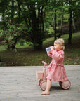 A young child in a pink dress sits barefoot on a pink tricycle on a wooden deck, sipping from a mushie Soft Spout Sippy Cup, with green trees and grass in the background.