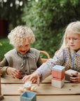 Two young children play outdoors at a wooden table, enjoying colorful wooden blocks, an abacus, and the mushie Suction Spinner Toy for a fun sensory experience. The lush green background suggests they are in a park or garden.