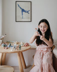 A young girl in a pink dress sits at a wooden table, playing with mushie Daisy Silicone Suction Builders and small figurines. Art supplies and bath toys are scattered nearby as sunlight highlights her motor skill development activities.