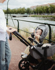 An adult in a white shirt and jeans strolls by the river with a baby holding a toy attached to the mushie Adjustable Silicone Strap 2-Pack, creating a peaceful scene among trees and grass.