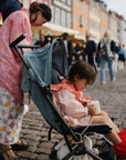 A young child sits in a stroller on a cobblestone street, holding a cup secured with the mushie Adjustable Silicone Strap 2-Pack. An adult stands nearby, while people and colorful buildings fill the lively street in the background.