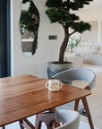 A mushie Snack Cup and a mug rest on a wooden dining table near two gray chairs in a bright, modern living space with a large potted plant, wall mirror, and neutral decor—perfect for toddlers.