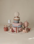 A selection of pastel mushie baby feeding items—including bowls, cups, utensils, a bottle, and Silicone Baby Food Containers—are neatly arranged on a round placemat against a beige background.