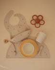 A set of beige and yellow baby feeding items, including a bib, suction bowl, spoon, Mushie Trainer Sippy Cup with easy-grip handles, pacifier with clip, and flower-shaped teether—each arranged on a matching placemat.