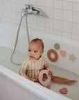 A toddler in a striped swimsuit sits in a shallow bathtub, holding a pink ring toy. mushie's Foam Bath Puzzle shapes stick to the tub’s side, making bath time both fun and educational.