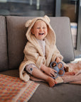 A happy toddler in a bear-ear bathrobe sits on a couch holding the mushie Daisy Rattle Teether. A patterned cloth and towel are nearby, and the child looks relaxed while playing with the teething toy indoors.