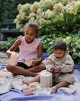 Two young children sit on a blanket outdoors, surrounded by greenery, enjoying pretend play with the Mushie Silicone Tea Play Set. One pours tea while the other examines a toy, with cups and a teapot from the set in front of them.