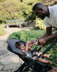 A man smiles as he hands a mushie Adjustable Silicone Strap 2-Pack to a baby in a stroller outdoors, surrounded by greenery, flowers, and a stone pathway.