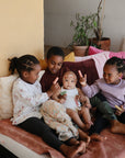 Four children sit on a bed with colorful cushions. Three older kids play with mushie Silicone Animal Finger Puppets to build fine motor skills, while a baby sits in the center. A potted plant appears in the background.
