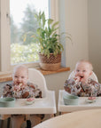 Two babies in matching mushie Long Sleeve Bibs sit in high chairs by a window, smiling as they snack from green bowls and cups. A potted plant sits on the windowsill behind them.