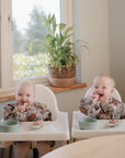 Two babies in matching mushie Long Sleeve Bibs sit in high chairs by a window, smiling as they snack from green bowls and cups. A potted plant sits on the windowsill behind them.