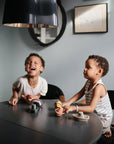 Two young children play at a dark table with mushie Bath Boats. A child in a white shirt laughs, while another in a striped tank top watches calmly. These non-toxic plastic boats bring fun to the softly lit, art-filled room.