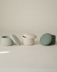 Three mushie Silicone Baby Food Containers in light green, cream, and sage are lined up on a neutral background; each small round food-grade silicone container has a ribbed lid resting slightly open or nearby.