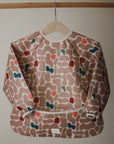 The mushie Long Sleeve Bib, featuring a water-resistant design and a colorful abstract pattern in pink, beige, blue, green, and red, hangs on a wooden hanger against a neutral background.
