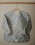 The mushie Long Sleeve Bib, featuring a blue floral print, hangs on a wooden hanger against a beige wall. This water-resistant, machine-washable bib has elastic cuffs and a wide pocket for catching spills.