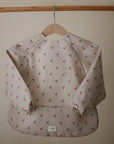 The mushie Long Sleeve Bib, featuring a light color and small cherry pattern, is water resistant and displayed on a wooden hanger against a plain beige background.