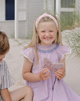 Girl in purple dress holds mushie Silicone Sensory Toy sitting on sand next to a boy.