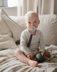 A baby sits on a couch wearing a striped onesie, holding a green toy and soothing with a FRIGG Silicone Pacifier from the FRIGG Silicone Pacifier 4-Pack, as sunlight streams in through the nearby window.