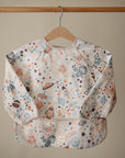 The mushie Long Sleeve Bib, water resistant and adorned with a space-themed print in orange, blue, and brown, is displayed on a wooden hanger against a beige background.