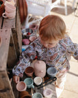 A toddler plays outdoors with the pastel-colored Silicone Tea Play Set by Mushie, wearing a floral top. Sunlight streams in, illuminating the scene surrounded by potted plants and a wooden shelf.