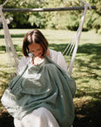 A woman sits on a hanging chair in a garden, smiling as she breastfeeds her baby under the mushie Muslin Nursing Cover. Sunlight streams through the trees, highlighting this serene moment.