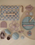 A selection of pastel mushie baby items—including a checkered pouch, utensils, divided plate, placemat, Daisy Rattle Teether, rainbow and boats toys, strawberry toy, and small geometric shapes—arranged neatly on a light background.