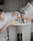 Two children sit at a small round table, playing with mushie Wooden Balance Blocks made from FSC-certified wood. They focus on building, while a white sofa and cushions appear in the background.