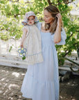 A woman in a light blue dress holds a young girl in a floral outfit and sun hat, who grips the mushie Daisy Rattle Teether. They stand outdoors on a sunny day among leafy greenery and dappled sunlight.