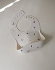 The mushie Silicone Baby Bib features an adjustable neck strap and playful animal and flower illustrations. Laid flat on a light surface, this easy-to-clean white bib is perfect for little ones.