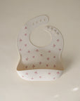 The mushie Silicone Baby Bib, made from non-toxic silicone, features small pink bow patterns and adjustable buttons, shown flat on a light beige background.