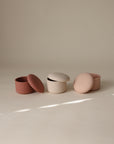 Three round, ribbed mushie Silicone Baby Food Containers in muted pink and beige rest on a neutral surface. Two matching dome lids are open, one is closed, and soft light creates gentle shadows.