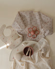 A flat lay of baby items in neutral and pastel tones with cherry prints, featuring the mushie Organic Cotton Muslin Swaddle Blanket, a long-sleeve smock, bib, pacifier clip, flower teether, bowl with utensils, and a soft toy ball.