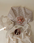 Flat lay of baby items in soft neutrals and cherry patterns, featuring the mushie Organic Cotton Muslin Swaddle Blanket along with a long-sleeve bib, flower teether, bowl and spoon set, textured ball, pacifier, and muslin cloth on a cream background.