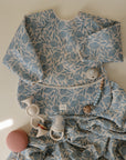A mushie Organic Cotton Muslin Swaddle Blanket and a baby bib with blue floral patterns are displayed on a light surface, accompanied by baby toys such as a rattle, teether, and a pink textured ball.