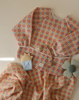 The mushie Organic Cotton Muslin Swaddle Blanket is displayed with colorful, patterned children’s clothing, an organic cotton pacifier clip, a soft teether, and a flower-shaped toy on a light beige background.