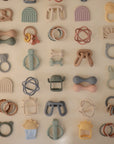 The mushie Rainbow Teething Mitten is a pastel-colored silicone mitten for babies, arranged alongside rattles in various animal, rainbow, circle, and geometric shapes on a light background.