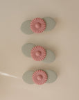Three light green oval-shaped mushie Suction Spinner Toys with pink daisy-like decorations are arranged vertically on a beige surface, offering a delightful sensory experience.