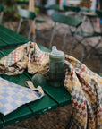 A green outdoor table with a checkered cloth holds a mushie Soft Spout Sippy Cup with a green lid, a blue and white checkered pouch, and a small ball. Green and wooden chairs can be seen in the background.