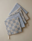 Four mushie Muslin Cotton Washcloths in soft blue and beige—some checkered, others solid—are layered on a light beige surface. Each washcloth has a small corner fabric loop for easy hanging.