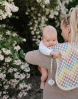 A woman stands near a bush with white flowers, holding a baby resting on a colorful checkered cloth. The baby uses a FRIGG Natural Rubber Pacifier 4-Pack, attached with a clip for comfort and style.