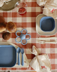 A table set for two children features mushie Square Dinnerware Plates—blue, BPA-free—with matching forks and spoons on a red-and-white checkered tablecloth, plus ribbon-tied napkins, bread rolls, a drink, and a blue flower-shaped teether.