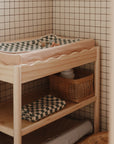 A wooden changing table with a green and white checkered diaper changing pad, Mushie Changing Pad Liners, a small toy on top, and wicker baskets for baby items on the lower shelves stands against a tiled wall.